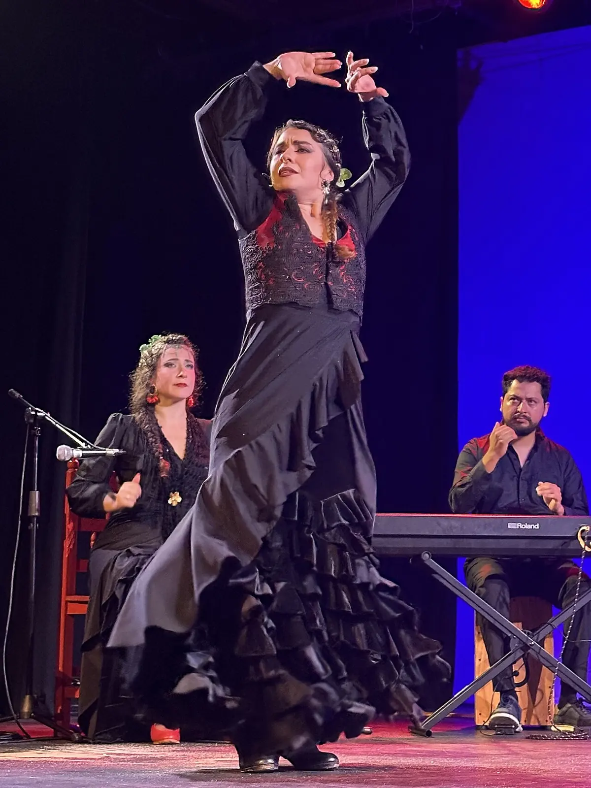 A flamenco dancer in a black ruffled dress raising her arms on a stage lit blue, with a singer seated at left and a keyboard player at right