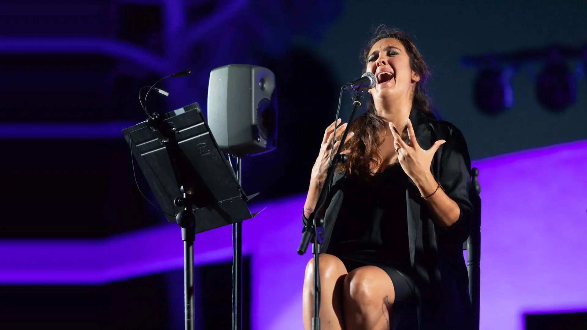 A flamenco singer seated on a stage with her hands raised, singing into a microphone under violet light